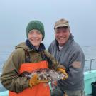 Two anglers standing on a boat, one of them holding a large brown and black lingcod.