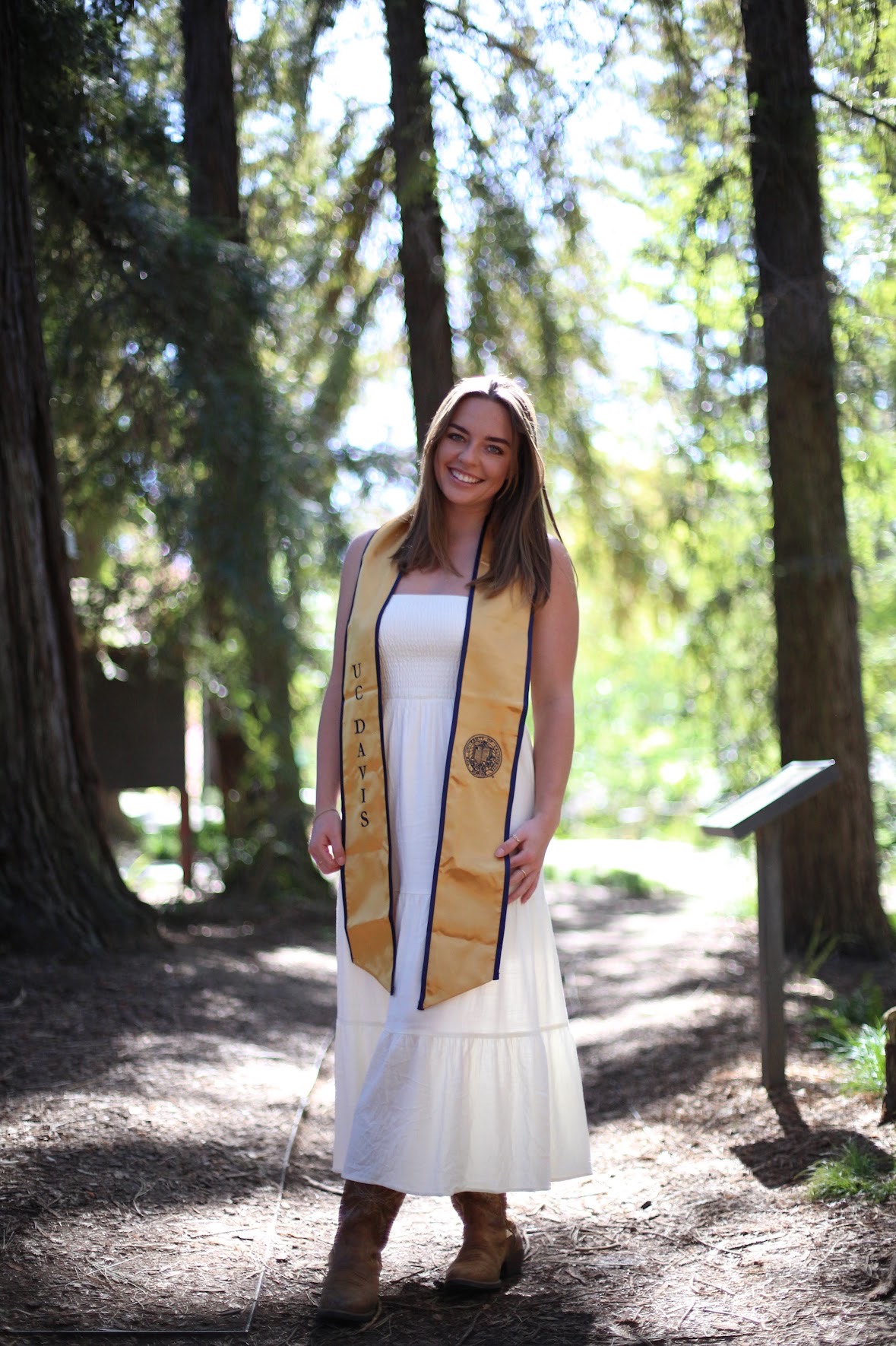 An image of a Maddie Wilmot standing in a redwood grove, wearing a white dress, boots, and a gold and blue UC Davis graduation shawl.