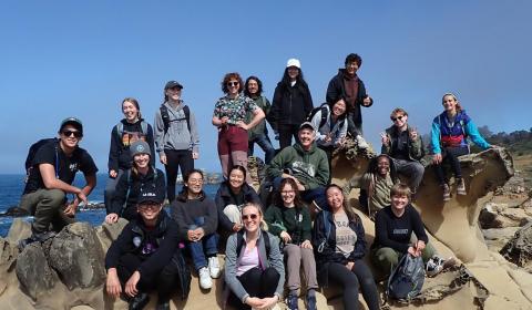 A group of students gathered on a rocky outcrop next to the ocean, smiling and posing for the photo.