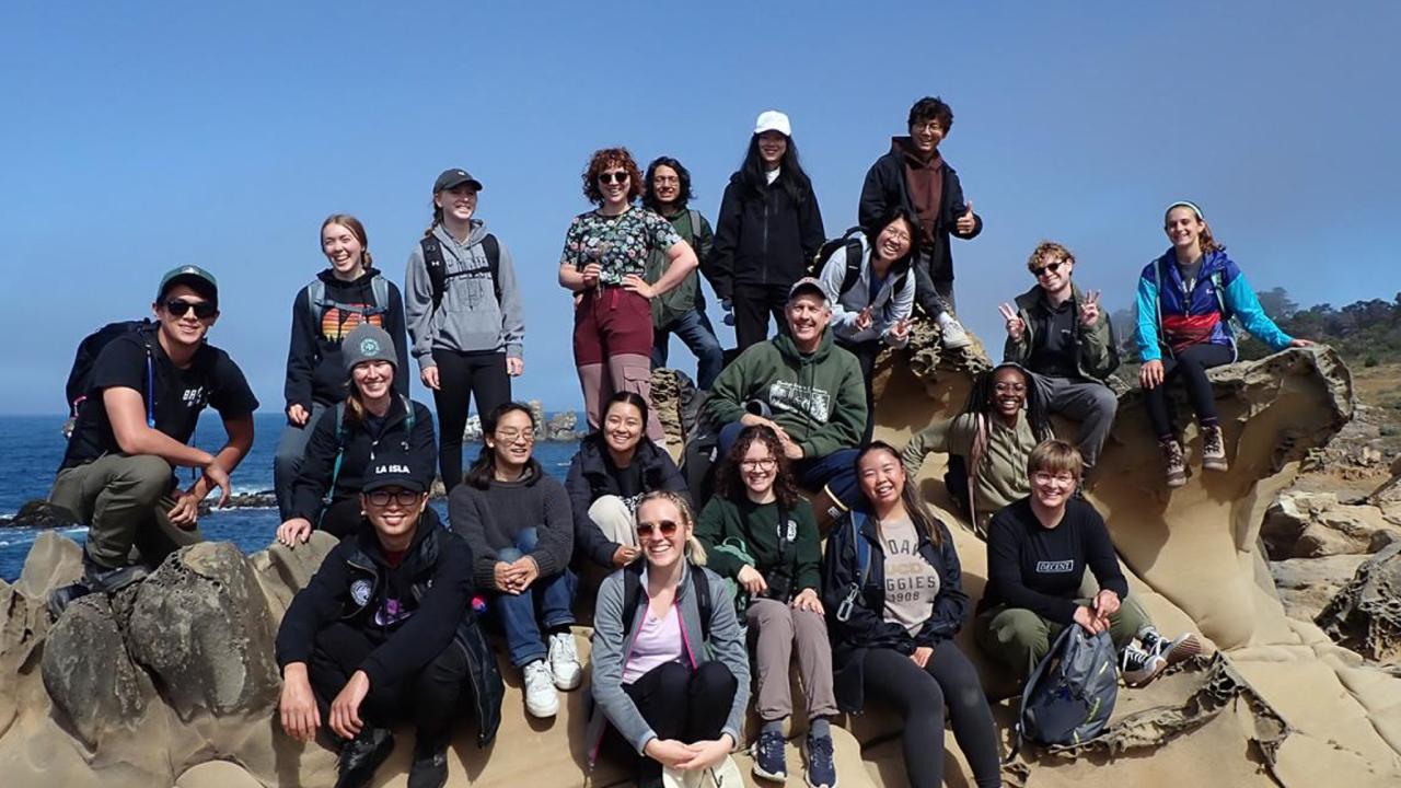 A group of students gathered on a rocky outcrop next to the ocean, smiling and posing for the photo.