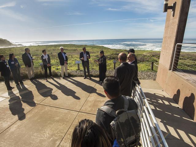 A group of people standing around watching one person present. They are outdoors, with a view of the coast and ocean in the background. Their shadows are stretching across the pavement because the sun is at their backs.