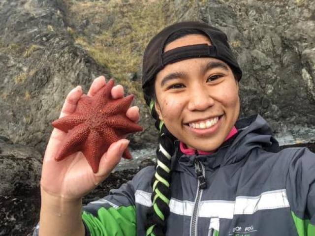 A person in a backwards baseball cap holding up a sea star next to their face