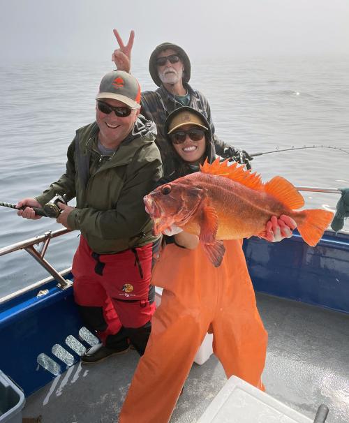 Three people standing together on a boat. One of them is holding up an enormous, bright orange rockfish.