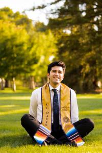 A person sitting cross legged on grass in front of trees, wearing a UC Davis graduation stole