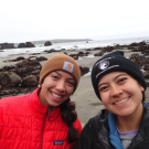 A close up of two people dressed for cold weather in beanies and jackets. Behind them is a rocky intertidal area.