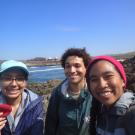 Three smiling students with the ocean in the background and the student on the left is holding up a hemoglobin plushie.