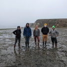 Five people posing and smiling in low tide mud flats behind oyster bags.